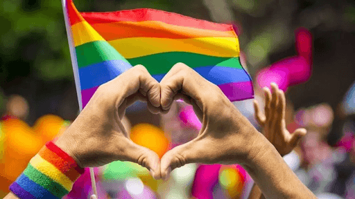 Hands forming a heart shape in front of a rainbow Pride flag at a LGBTQ+ Pride celebration, with a rainbow wristband and festive background.
